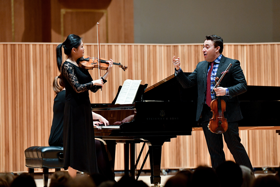 A female Asian student performing the violin, with Maxim Venegerov, a white male violinist, teaches her, with a female pianist performing alongside the student in the background.